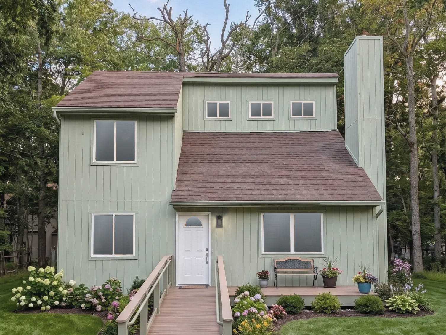 A two-story house with sage-green siding among tall trees, with a wooden ramp to the front door and flower beds along the front.